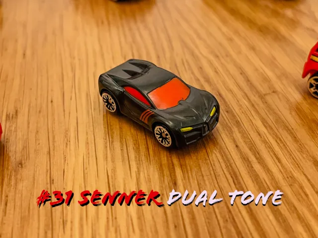 A carousel photo displaying the thirty-first vehicle named 'Senner Dual Tone' out of an array of 'Deluxe Cars', at full size. The car is painted mostly black with the following components orange; windshield and sides. The front-lights are painted yellow.