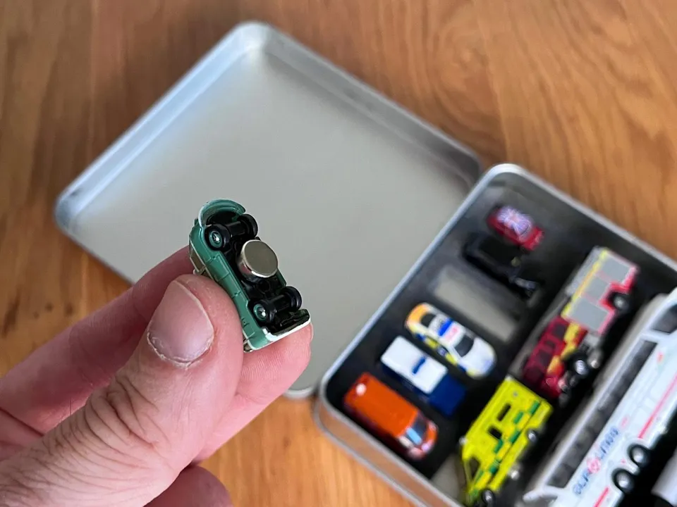 Julian holding a green blue magnetic bus via three fingers, whilst in the background you see a up close shot of the V.I.A. metal tin comprise of the other vehicles.